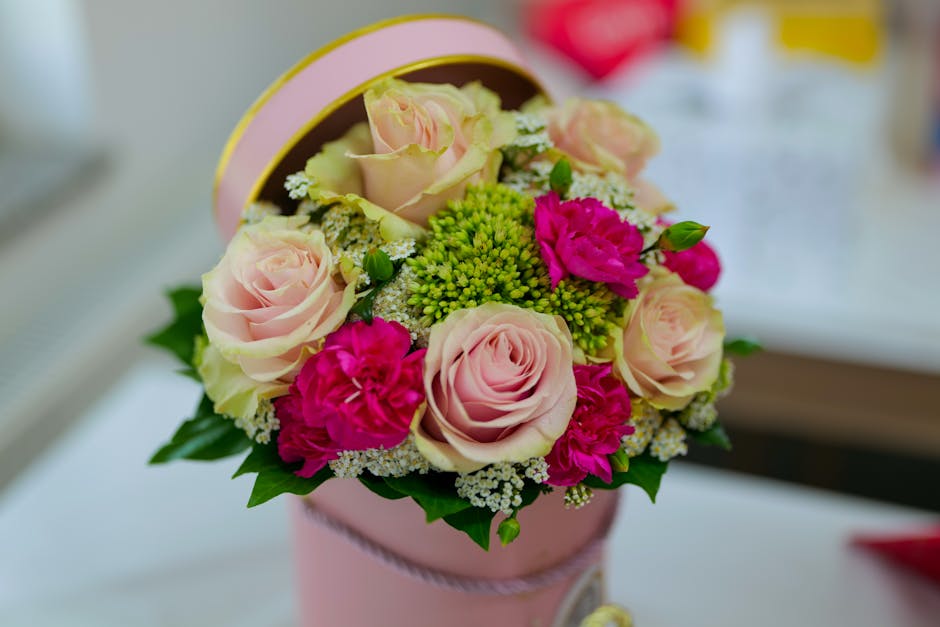 A round, pink floral arrangement featuring large, soft blush roses with gently unfurling petals, complemented by vibrant magenta carnations and clusters of small white and lime green filler flowers. T
