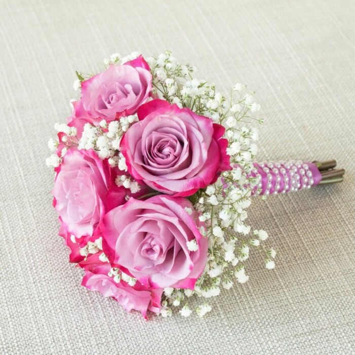 An exquisite, hand-tied wedding bouquet inspired by Epping's rustic charm and rich floral heritage, this arrangement features five prominent roses whose petals seamlessly blend from deep fuchsia at the tips to soft lavender at the centre. Each bloom is artfully cradled among sprigs of baby's breath, their tiny, white florets adding a frothy delicacy that contrasts delightfully with the roses' velvety texture. The flowers are bound with a satin ribbon in regal purple, its smooth sheen adorned by parallel rows of luminous pearls woven in a spiral-an elegant nod to the classic English bridal aesthetic. The bouquet's natural green stems peek from below, carefully trimmed to enhance the arrangement's tidiness and organic feel. Resting on a textured, pale-beige cloth, the bouquet's rounded silhouette and sophisticated palette are highlighted by soft, diffused daylight-recalling the gentle ambiance of a wedding morning in Epping's countryside. The arrangement, seen from above, not only displays the intricate work of a skilled florist but also evokes emotional resonance: it captures the hope, anticipation, and joy of a new chapter as it radiates cheer and tenderness. Perfect as a bridesmaid or flower girl bouquet or as a refined centrepiece for spring and summer festivities in Essex, it's a visual tribute to enduring romance, meticulous artisanship, and the picturesque landscapes surrounding Epping.