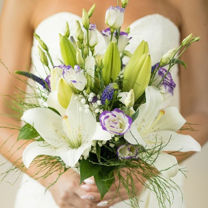 In this radiant close-up, a bride framed by the subtle light of Epping's countryside cradles a bouquet that epitomizes both classic and contemporary elegance. Dominating the arrangement are large, snowy white Oriental lilies at their peak bloom, some still unfurling their pale, sculptural petals, each speckled subtly and offset by bright, pollen-laden stamens. Interspaced with care are clusters of delicate lisianthus, their ruffled white petals edged with a whisper-thin band of lavender, adding a hint of unexpected colour-an artistic echo of local bluebells found in Epping Forest. Deeper indigo Veronica, bold in form and hue, lend sophistication and visual balance, while puffs of baby's breath float between, creating a soft, lace-like texture that diffuses the ensemble. The arrangement's vigor is amplified by abundant fresh green foliage: broad, glossy leaves, unopened lily buds bursting with promise, and airy asparagus fern encircle the composition. The bride's relaxed hands, her nails painted a gentle pink, suggest quiet confidence, their position at the bouquet's base bringing a sense of movement and grace. In the blurred background, her strapless ivory gown lends an ethereal radiance, highlighting the bouquet as the story's centerpiece. Perfect for wedding inspiration on Epping florist blogs, this bouquet resonates with the romance and natural beauty of Essex, making it an unmistakable choice for modern couples seeking a wedding infused with local charm and timeless artistry.