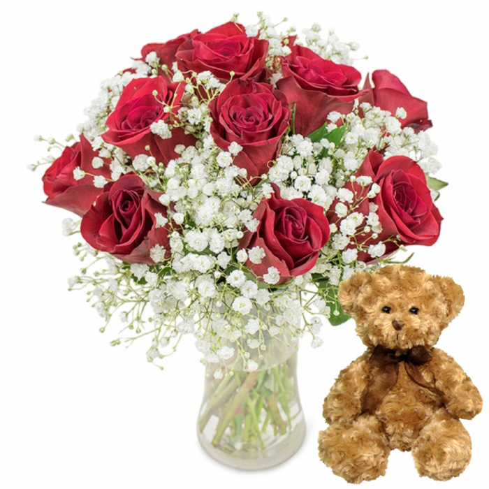 The photograph presents a beautifully composed romantic gift set, styled as though it is ready for same-day delivery to homes and offices across Epping and the surrounding villages. On the left, a tall, clear glass vase holds a sumptuous bouquet of roughly one dozen deep crimson red roses. The roses are at various stages of opening, yet each bloom looks perfectly formed, with velvety petals spiralling outward in tight, symmetrical layers that catch the light and create subtle shadows. Their rich, wine-toned red hue is instantly evocative of love and devotion. Interlaced with the roses are abundant stems of white baby's breath, the delicate clusters of miniature flowers forming a fine, misty backdrop that softens the intense reds and adds volume and a sense of airiness to the arrangement. Through the transparent glass, straight green stems are visible, neatly trimmed and resting in clear water, emphasizing freshness and professional care that might remind a viewer of stepping into a florist just off Epping High Street. To the right of the vase, a soft, light brown teddy bear sits attentively, its plush fur looking fluffy and inviting to the touch. The bear's rounded ears, small black button eyes, and gently embroidered nose and smile give it a warm, reassuring expression, like a quiet companion to the roses. A dark brown satin bow is tied snugly around its neck, its sheen providing a subtle contrast to the matte fur. Together, the bouquet and teddy bear create a scene of thoughtful romance, ideal for marking anniversaries, Valentine's Day, or heartfelt apologies for couples living near Epping Forest, Theydon Bois, or Coopersale.