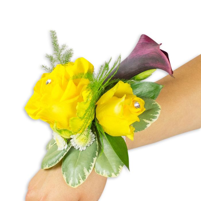 An expertly arranged wrist corsage lies diagonally across a light-toned forearm on a pristine white background, composed to showcase both floral detail and wearable elegance. The focal pair of yellow roses - the left blossom fully opened, soft and sunlit, the right one smaller and more cupped - are set with discreet clear rhinestones at their centres so they catch light like tiny, celebratory sparks. Behind them, a single deep burgundy-to-plum calla lily rises with a smooth, sculptural curve, its dark interior crafting a refined contrast against the warm yellow. Around the flowers a variety of greenery adds depth and texture: fine, fern-like asparagus shoots create a soft halo and gentle movement; broader variegated leaves with pale margins offer weight and glossy, cooling surfaces; and small white button fillers tuck into the composition to soften edges and suggest fullness. The arrangement sits on the wrist with a slim, mostly hidden band, demonstrating how such a corsage stays comfortably in place for long services or evenings out. As a piece intended for weddings, prom nights, or distinguished special occasions, this design channels the optimism of yellow with the formality of deep purple - a palette that photographs well beneath the trees of Epping Forest or at intimate gatherings along Station Road, and that reflects careful handcraft from a local artisan florist familiar with the town's tastes.