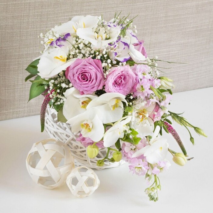 A refined floral centerpiece is presented in a rounded white wicker basket, photographed in a way that makes every bloom appear crisp and freshly arranged, as if ready to be placed on a wedding table at a country venue just outside Epping. In the centre of the composition, several large pink roses, fully opened, reveal intricate spirals of petals in tones from soft blush to slightly deeper rose, suggesting tenderness and romance. Surrounding and weaving through these roses are elegant white Phalaenopsis orchids, their broad, smooth petals catching the light, while their yellow-orange centres with subtle markings add an exotic, high-end detail often chosen by local couples planning celebrations near Epping Forest. Delicate Lisianthus flowers in white, some with edges brushed in pale lavender, nestle between the larger blossoms, contributing a ruffled softness and hint of cool purple to the predominantly pink-and-white palette. Stretching outward, slender stems of pale pink stock cascade gently over the basket's edge, their many small florets creating a feathery texture and vertical interest, as if the flowers were just gathered from a well-kept garden in Coopersale. Fine sprays of white baby's breath are lightly tucked throughout the design, softening transitions and filling the spaces with a mist-like lightness that makes the arrangement feel cloud-soft and airy. Fresh green foliage, in varying shades from deep to bright, peeks out between the blooms, adding structure and a natural, garden-fresh feel. The white wicker basket with its intricate weave rests on a spotless white surface, accompanied by two decorative woven balls made from pale natural fibres that echo the basket's texture. The neutral, linen-textured beige backdrop stays quietly in the background, allowing the carefully balanced colours and thoughtful florist craftsmanship to stand out, ideal for elegant events and home styling throughout the Epping area.
