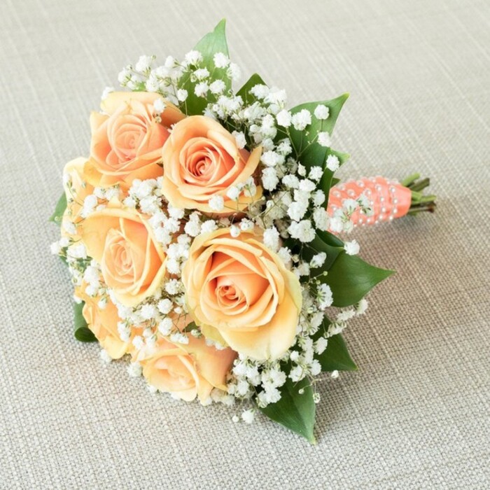 A gracefully composed bouquet of peach roses and white baby's breath rests horizontally across a lightly textured, linen-like beige surface, resembling the neutral linens often used on wedding tables in Epping venues. The arrangement is compact and round, tilting slightly towards the upper right of the image, as though just set down by a bride pausing for a moment of calm. At its heart, around seven to eight roses are gathered closely together, each bloom fully open or on the verge of unfurling. Their petals form intricate spirals, moving from tight, warm centres to fuller, softly curled edges in a palette of warm peach, soft apricot, and pale orange. These velvety petals seem delicate yet full, conveying both freshness and a sense of careful preparation by an experienced florist. Surrounding and weaving through these roses are abundant clusters of tiny white baby's breath, or gypsophila, creating an ethereal, cloud-like halo. The gypsophila's myriad miniature blooms offer a crisp white contrast and lend a light, airy texture that softens the structured round form. Interspersed are small glimpses of rich, green rose foliage, each leaf smooth and slightly glossy, providing depth and a natural, garden-fresh feel reminiscent of blooms gathered from homes around the Epping and Loughton area. The stems are neatly aligned and wrapped in a coral-peach satin ribbon, the colour chosen to echo the roses and harmonise with their warmth. Along this ribbon, small round white faux pearls are carefully positioned in a delicate spiral pattern, adding a sophisticated bridal detail often requested by couples planning local ceremonies near Epping Forest. The very ends of the stems are freshly cut and visible at the base, hinting at the bouquet's hand-tied craftsmanship. Soft, even lighting brings out the gentle shadows between the petals and highlights the pearly accents, giving the entire bouquet a serene, romantic presence ideal for weddings, bridal showers, or engagement celebrations in and around Epping.