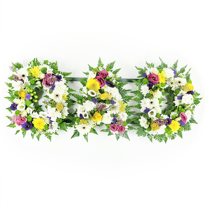 Displayed against a clean white studio background, this photograph captures a classic DAD floral tribute, the kind families in Epping often choose when they want something both traditional and personal for a father's funeral. Three bold capital letters - D, A, and D - are arranged in a horizontal line, each letter built upon a sturdy frame softened completely by foliage and blooms. Around the outer edges, cascading green fern leaves curl gently, creating a natural, feathery border that defines the contours of each character. The interior of the letters is packed tightly with pristine white chrysanthemums and generous white gerbera daisies, their soft, rounded petals overlapping like layers of fabric, evoking purity, remembrance, and peace. Threaded through this white base, bright yellow gerbera heads and sprigs of yellow spray roses appear in balanced clusters, offering little flashes of optimism and gratitude, like sunlight touching a quiet woodland clearing near Epping Forest. Deeper shades of purple lisianthus and rich fuchsia or magenta spray roses nestle between the lighter blooms, providing contrast and emotional depth, as if to acknowledge both loss and enduring love. Small lavender flowers and green button chrysanthemums are dotted throughout, adding fine texture and a gentle contemporary note. All the flowers are arranged front-facing, with careful spacing so that every letter remains clearly readable from the front pew of a chapel or from across a graveside. Discreet dark support bars hold the letters together, almost invisible beneath the foliage, uniting them into one coherent message. The tribute feels lovingly considered, the kind of arrangement a local florist near Epping High Street might create to help a family express everything they wish they could say to their dad in one beautiful, fragrant gesture.