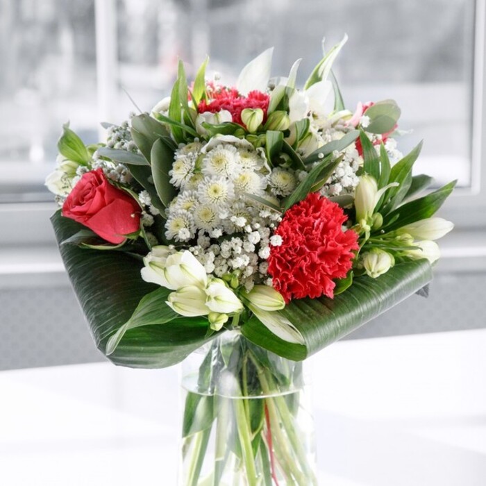 A refined yet exuberant floral arrangement commands the centre of the image, poised in a sleek cylindrical glass vase whose transparency reveals the tidy bundle of green stems beneath the water. The bouquet itself is a dance between passionate reds and serene whites, very much the sort of design you might find gracing a dining table in an Epping home after a special same-day delivery. Velvety deep red roses form the romantic core, their petals richly layered and just beginning to unfurl, while a single large crimson carnation at the front adds intricate, ruffled texture that feels almost like carefully gathered silk. Nestled among these are white alstroemeria, their slender petals opening with quiet elegance and dusted inside with faint speckling, along with plump white lily buds held upright like graceful promises of tomorrow's blossoms. Tiny clusters of gypsophila scatter through the arrangement, each minuscule white flower adding a sense of lightness, as if a soft flurry of snow had settled gently between the more dramatic blooms. Framing everything is an abundance of green foliage: wide, glossy dark leaves are wrapped and tucked around the base, creating a sculpted collar that feels modern and polished, while narrower leaves and eucalyptus-like sprigs stretch upward, introducing fresh shades of green and a slightly wild, garden-like rhythm. The bouquet rests on a spotless white surface against a bright, softly out-of-focus backdrop, the kind of light you might see pouring through a window overlooking Epping's leafy streets near Bell Common. The mood is both celebratory and composed-a bouquet equally suited to taking pride of place at a family gathering, offering gratitude to a host, or brightening an office just off Epping High Street with a sense of thoughtful care and enduring affection.