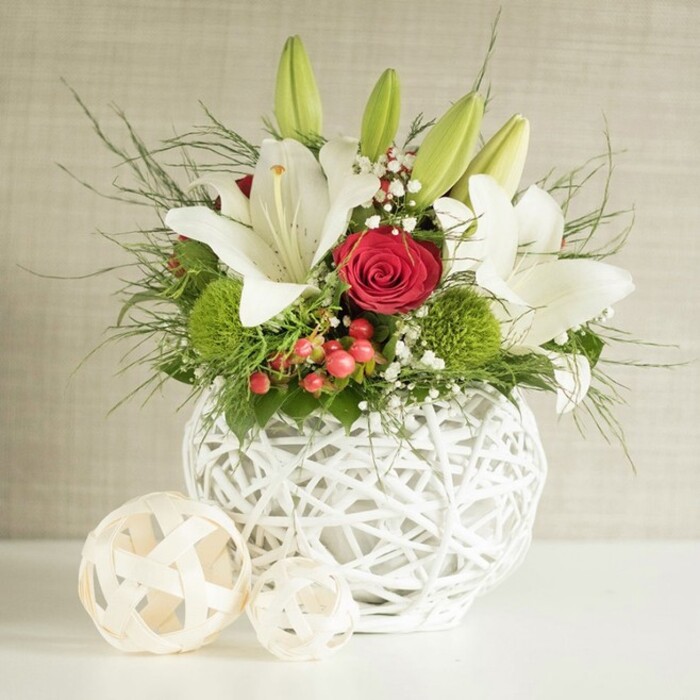 On a pristine white tabletop, a refined floral arrangement is staged as if ready for a wedding reception at a boutique hotel near Epping station. The focal point is a striking, round white vase, fashioned from intertwining strands that form an open, basket-like structure with plenty of negative space, allowing soft shadows to play across the surface. Rising from this artistic container is a lush composition dominated by white lilies, their star-shaped blooms unfolding into generous, satin-like petals. Some lilies are fully open, showcasing tall yellow stamens and a hint of speckling near the throats, while others remain as smooth, tapered green buds, adding height and anticipation to the design. Tucked thoughtfully among them are deep red roses, one particularly prominent bloom at the center, its spiralled petals opening in tight, precise layers that bring a sense of romance and devotion often associated with wedding or anniversary flowers in the Epping area. Vivid green pom-pom flowers, likely chrysanthemums or dianthus, appear like soft mossy orbs, introducing a playful textural contrast and a refreshing splash of colour. Delicate sprigs of baby's breath float between the larger flowers, creating a mist-like softness, while clusters of small red berries lend warmth and seasonal charm that would suit a winter table at a local Epping venue. Wisps of fine, feathery foliage-resembling asparagus fern-trail outward, mingling with darker, broader leaves that provide depth and a natural framework. To the left of the vase, two decorative woven spheres in pale, natural tones rest casually, their open-lattice pattern mirroring the vase and tying the whole composition together. The neutral beige background keeps the focus on the vivid reds, pure whites, and lush greens, capturing the mood of quiet celebration crafted with the skill and eye of a local Epping florist.
