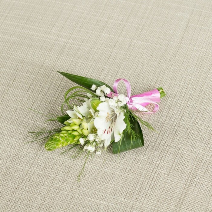 An elegant, compact boutonniere is displayed against a softly textured, light beige background that resembles fine linen, its muted tone providing a calm, neutral stage for the flowers to shine. At the centre of the composition is a single white alstroemeria blossom, also known as a Peruvian lily, its petals broad and gently curving, with tiny reddish-brown flecks concentrated near the throat that lend a painterly intricacy. Nestled alongside this main bloom is a conical cluster of vivid lime-green buds, each tightly packed to form a tapered spike that adds a modern, sculptural element to the design. Sprinkled throughout are small sprays of white baby's breath, their cloud-like clusters forming a delicate veil that softens the strong shapes of the main flowers and gives the arrangement a light, romantic air. Supporting these blooms is a gathering of dark green, sword-like leaves, their pointed tips fanning out to frame the boutonniere, while thinner, grass-like strands curl and loop, introducing texture and a sense of lively movement. The stems are neatly bound on one side with a smooth, bright pink satin ribbon, tied into a precise little bow whose cheerful sheen contrasts beautifully with the natural fibres of the background. Gentle, even lighting washes over the scene, catching the satin's lustre and the alstroemeria's subtle petal veining without harsh highlights, creating a serene, polished look. This small floral piece would be perfectly at home on the lapel of a groom at an Epping wedding venue or a guest attending a formal event near Epping Forest, evoking themes of freshness, tenderness, and thoughtful local craftsmanship in every detail.
