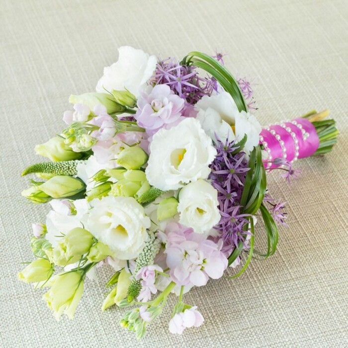 Gently placed at a refined diagonal atop light beige linen, this vibrant Epping-inspired bouquet melds classic romance with contemporary flair, drawing together creamy white Eustoma as its star. Their ruffled petals-the softest ivory tipped in pale pastel yellow-open wide beside slender, lime-green buds, symbolising possibility and fresh beginnings. Interwoven among them are pastel lavender-pink stock, their clouds of petals adding impressions of fragrance and plush texture evocative of early spring blooms found along Epping's greenways. The striking geometry of deep purple allium, their star-like clusters radiating structure and drama, elevates the arrangement with unforgettable presence. Flecks of verdant foliage, including looping blades of bear grass and fuzzy green fillers, create a dynamic foundation that echoes the wild yet refined beauty of local meadows. A sweep of bold fuchsia satin ribbon binds the stems, finished with a luminous strand of white pearls-an elegant nod to wedding-day tradition and celebration, reminiscent of the charm in local venues like Copped Hall. Soft, diffused lighting enhances every nuanced color and detail, inviting admiration for the floristry craft. This bouquet perfectly embodies the festive yet sophisticated spirit of Epping weddings, making it a timeless choice for local brides, bridesmaids, or special celebrations that yearn for romance, artistry, and natural grace.