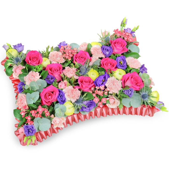 A carefully constructed pillow-shaped memorial arrangement, photographed slightly from above so the dense surface reads like a soft, floral textile, communicates both craft and compassion. The central plane is dominated by numerous hot pink roses, their petals velvety and full, creating a rich, tactile focal area surrounded by sprays of pale pink carnations whose frilly edges give a gentle, cloud-like texture. Interspersed throughout are deep purple lisianthus for tonal contrast and bright lemon-yellow ranunculus that inject cheerful highlights, together composing a lively, life-affirming palette. Cool accents of silver-blue eryngium thistle are positioned to catch the eye with their spiky form and metallic tone, adding structure and a hint of wild beauty. Sprays of rounded eucalyptus leaves and varied green fillers provide depth and a silvered-green backdrop that allows the colours to sing while offering a subtle fragrance. The whole composition is seated upon a ruffled, pleated red satin base that neatly defines the pillow silhouette and adds a formal, satin sheen around the perimeter. The soft, diffused lighting flatters the petals and ribbon without dramatic shadows, creating an atmosphere of quiet dignity and gentle solace. This is a traditional funeral pillow-vibrant yet tender-designed to celebrate a life with warmth rather than dark mourning; its fuchsia, violet, lemon and blush tones convey affection, remembrance and comfort for family and friends during services, memorial displays or graveside tributes across the Epping area.