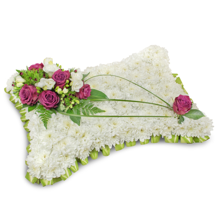 A classic pillow-shaped funeral tribute sits on a stark white background, its form defined by a dense surface of immaculate white chrysanthemum florets that read as a soft, cloudlike cushion. Each small chrysanthemum is placed closely to achieve a continuous, plush texture that evokes serenity and repose - an intentionally crafted foundation for a sympathy piece. In the top-left corner a rich cluster of deep fuchsia or magenta roses forms the primary accent: velvety, fully opened heads juxtaposed with delicate white freesia or lisianthus that introduce lightness, scent, and a whisper of fragility. Surrounding the flowers, an abundance of fresh green foliage - including feathery fern fronds and glossy leaves - creates a verdant backdrop that heightens the contrast with the pure whites and deep pinks. Across the main field of the pillow three slender stems of dark green bear grass arch gracefully, drawing an elegant diagonal line that guides the eye from the floral cluster toward the opposing corner, where a single matching magenta rose is placed with mindful balance. The entire perimeter is finished with a folded lime-green satin ribbon, forming a neat scalloped trim that frames the design and adds a subtle, uplifting colour edge. Photographed in soft, even light so textures and colours read clearly, this arrangement communicates solemnity, comfort, and respectful remembrance. As local florists in Epping often advise families, this pillow is a traditional expression of condolence suited to funeral chapels near Epping High Street or memorials beside Epping Forest, and is the sort of thoughtful tribute we craft at Flowers Epping with careful attention to proportion and sentiment.