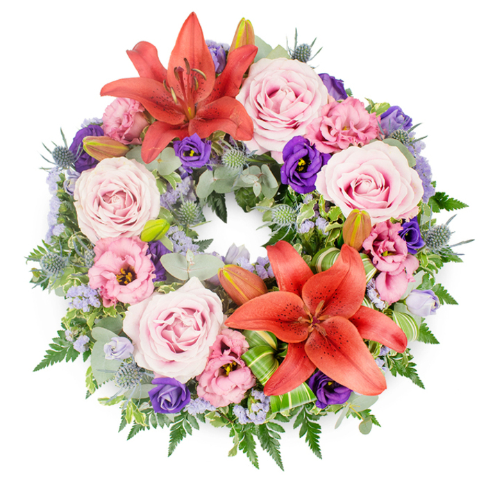 Captured from overhead on a clean white backdrop, this meticulously crafted funeral wreath forms a complete circle, symbolising eternity and the ongoing cycle of love and remembrance. The ring is densely filled, giving a sense of generosity and care, with no gaps in the floral coverage. Dominating the composition are two large coral-orange Asiatic lilies, their trumpet-shaped blooms fully open, each petal gently arched and lightly speckled, with central stamens tipped in deeper rust tones. One lily sits near the upper arc of the wreath, the other slightly off-centre toward the lower side, creating a natural, flowing visual rhythm. Scattered between these focal flowers are numerous roses in shades of blush, soft pink and lavender-pink, their tightly layered petals unfurling into rounded, romantic blooms that convey tenderness and sympathy. Mixed through the roses are lisianthus (eustoma) blossoms, some a gentle pastel pink, others a rich, velvety purple, their ruffled petals adding a sense of movement and sophistication. Fine clusters of tiny lavender-blue filler flowers, likely statice or limonium, drift through the design like a soft mist, softening transitions and echoing the cool tones often seen in early morning light over Epping's open fields. The foundation of the wreath is a varied bed of foliage: silvery eucalyptus leaves, airy fern fronds, and other deep green leaves weave together to provide structure and depth, while spiky blue-grey eryngium thistles introduce a striking, sculptural contrast. Several closed lily buds appear discreetly among the greenery, hinting at life still unfolding. Near the bottom section, folded, glossy green leaves-similar to aspidistra or ti leaves-are arranged with a contemporary flair, showing the hand of a skilled local florist accustomed to creating tributes for services at Epping churches and the nearby crematorium. The overall effect is peaceful and dignified, an expression of respect, comfort, and enduring memory.