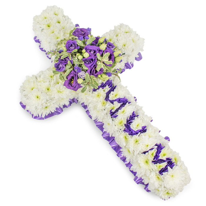 This striking funeral cross is composed of a dense layer of pristine white chrysanthemums that create a soft, pillowy foundation, each bloom deliberately set to form a continuous, serene surface. Against a pure white backdrop the white chrysanthemums read as a field of light, their full petals diffusing the soft studio illumination and reinforcing a mood of tranquility. A deep purple satin ribbon, pleated along the outer edge, provides a solemn border; its ruffle outlines the cross with classical refinement and lends a quiet contrast between the cool whites and rich purples.
At the crossroads of the arms, a lush, rounded spray becomes the arrangement's centrepiece. Deep violet roses, their petals compact and velvety, sit beside lighter lavender roses and ruffled eustoma (lisianthus), the tonal shifts from duskier violets to gentle lavenders creating a layered, contemplative effect. Variegated foliage-leaves brushed with cream at the margins-adds an architectural relief, while small green berries are interspersed to give the cluster an organic, tactile accent. On the vertical arm, the name MUM is crafted from individual purple flowerheads, the letters raised slightly above the chrysanthemum base so the tribute reads as both personal and reverent.
Imagined for use at a funeral service, wake or graveside in the Epping area-perhaps laid near St. John's or at a family home off The Green-this piece balances dignity and tenderness. Its cool, layered palette suggests both purity and mourning, and the careful craftsmanship communicates enduring respect and heartfelt remembrance.