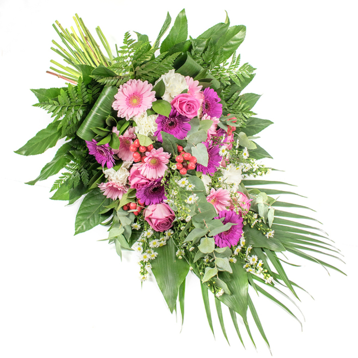 This striking diagonal funeral spray, photographed against a crisp white backdrop, appears almost as if it is gently drifting from the upper left to the lower right, guiding the eye in a soft, flowing line. At its heart are generous gerbera daisies, some a pale, blush pink and others a richer fuchsia and magenta, their round, open faces acting as joyful yet tender focal points. Around them, elegant pink roses, with their classic spiralled centres and softly cupped petals, speak of love, gratitude, and remembrance. Ruffled white blooms-most likely mini carnations or small chrysanthemums-are nestled throughout, adding both volume and a cloud-like brightness that contrasts beautifully with the stronger colours. Threaded between these larger flowers are delicate white daisy-style fillers, evoking chamomile or Matricaria, bringing a sense of lightness and calm, like wildflowers that might edge the paths around Epping's green spaces. Bright red hypericum berries appear in subtle clusters, their polished surfaces catching the light and adding a textural accent that hints at life and continuity. All of this colour is supported by an abundance of foliage: glossy, dark green leaves form a sturdy base; feathery fern fronds soften the outline; and rounded, silver-green eucalyptus adds a modern, fragrant touch. From the lower right, long, narrow palm leaves fan outward, enhancing the sense of movement and creating a gentle cascade. At the upper left, the bound stems are clearly visible, underscoring the hand-crafted nature of the piece. Designed as a casket spray or prominent tribute for a funeral service in Epping, this arrangement conveys deep sympathy, peace, and a heartfelt farewell.
