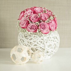 A romantic floral still life captures a domed bouquet of soft pink roses cradled in a contemporary white woven vase, the kind of arrangement you might find brightening a sunlit kitchen in a period home near Epping Forest. The roses are tightly massed yet individually distinct, each head opening into generous layers of velvety petals that transition from a pale, almost porcelain pink at the centre to a richer, deeper pink at the outer edges. Between the roses float clusters of tiny, star-like white baby's breath, their airy sprays softening the profile of the bouquet and lending it a dreamy, clouded halo effect. The vase itself is a globe of white-painted, intertwined fibres, with an open lattice design that gives the piece a handmade, artisan character while keeping the overall look clean and modern. It stands on a smooth white surface that gently mirrors the stems and base, suggesting a tidy console table or sideboard in a carefully curated Epping home. To the side and slightly in front of the vase sit two light beige decorative spheres crafted from narrow, woven wooden bands, their delicate, cage-like structure echoing the texture of the vase and contributing to a cohesive, nature-inspired arrangement. The background is a calm, warm beige wall, softly textured and evenly lit, allowing the pinks and whites of the flowers to glow without distraction. The scene carries a sense of quiet celebration and tenderness, ideal for marking intimate milestones, from a birthday breakfast in Theydon Bois to a simple thinking of you gesture delivered to a friend living off Station Road.