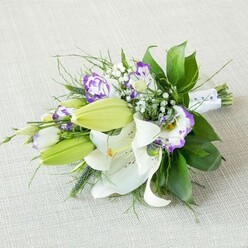An elegant, elongated bouquet lies on a pale, textured fabric that looks a little like finely woven linen, its neutral beige tone making the flowers appear even crisper and fresher. Dominating the composition are several open white lilies, their broad petals slightly reflexed at the tips and patterned with subtle speckles, each bloom presenting a cluster of yellow-orange stamens at the centre. Close by, a few bright green lily buds stretch forward, still smooth and closed, adding a sense of anticipation and growth. Woven among these lilies are Eustoma, or Lisianthus, with ruffled, cup-shaped blossoms in creamy white and striking violet-purple rims, some petals washed with softer lavender shades that suggest gentle movement and softness. Delicate sprays of baby's breath are scattered throughout, their tiny white flowers creating a misty, cloud-like veil that lightens the whole design, reminiscent of early-morning mist over the fields just outside Epping. Lush greenery anchors the arrangement, from broad, glossy dark green leaves that form a supportive base, to finer, more feathery foliage and a few spikier elements that add a modern, textural twist. At the narrower end of the bouquet, the stems are bundled neatly and wrapped in a smooth white satin ribbon, the surface dotted with small pearl-like beads that evoke the detail of bridal gowns seen at weddings around Epping High Street churches. The lighting is soft and even, with no harsh shadows, gently illuminating the white petals and making the purples appear rich yet calm. The bouquet feels both contemporary and timeless, ideal as a bridal bouquet, a special anniversary gift, or an elegant centerpiece for a dinner party at home in Epping.