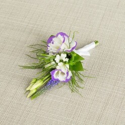 A single, elegant boutonniere lies carefully placed on a pale beige linen cloth, the fabric's faint weave giving the image a soft, tactile depth without taking attention away from the flowers. Two main lisianthus blossoms form the focal point, their white petals opening in generous, rose-like layers, each ruffled edge dipped in a rich, velvety purple that catches the light and draws the eye. Between them, miniature white filler flowers cluster like tiny stars, adding a light, airy texture that contrasts beautifully with the smooth lisianthus petals. Rising just behind is a slim spike of muscari, its tightly packed, deep purple bells introducing an additional note of colour and a subtle woodland feel, not unlike the wild blooms you might glimpse along the lanes leading out of Epping towards Theydon Bois. A couple of pale green buds rest alongside, still closed, suggesting that the arrangement was prepared fresh that morning, ready for a ceremony or prom night. Feathery green foliage, likely asparagus fern, spills gently around the blooms, while slightly broader, darker leaves provide structure, framing the flowers without overwhelming them. The stems are neatly aligned and bound in a band of white satin ribbon, the wrap smooth and slightly lustrous, finished with a delicate bow that signals the care of an experienced florist's hand. The overall feel is calm and romantic, perfect for a groom stepping out from a townhouse near Epping High Street or a guest arriving at a local country venue, the buttonhole offering a quiet accent of purple and white that speaks of thoughtfulness and understated style.