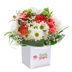 Evoking the soft, hopeful energy of a new day in Epping, this luxurious bouquet is cradled within a polished white gift bag that sits upright, ready to delight. The arrangement is a masterful blend of textures and tones: lush carnation blooms in shades from peach to vibrant red unfurl gracefully, each petal boasting intricate ruffles and a touchable softness. Nestled among them, crisp white daisies with glowing lime-yellow centers reflect a cheerful optimism reminiscent of a stroll along Epping's market streets. Airy sprays of baby's breath meander through the bouquet, lending a feathery lightness that invites lingering gazes, while hints of purple-tinged buds on the left edge add mystery and subtle sophistication. Rich, glossy green leaves surround the base, forming a protective embrace that gives the composition cohesion and elegance-the mark of an attentive florist's hand. The bouquet's shape is rounded and generous, a physical interpretation of abundance and celebration. Set against a clean white background, the arrangement's form and colour spring to life, making it not only a floral gift but a joyful occasion in itself. Apt for any hello, happy birthday, or expression of gratitude across Epping, this bouquet promises delight and a touch of local charm with every detail observed and every petal in place.