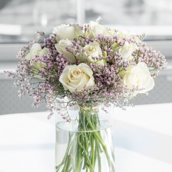 This image shows an elegantly composed sympathy bouquet that feels as peaceful as an early walk along the fringes of Epping Forest. Dominating the scene are generous white and creamy-ivory roses, fully open so that each blossom reveals a harmonious spiral of petals, curving inward like a quiet breath. Their surfaces appear satiny and smooth, catching the soft ambient light that falls from one side of the frame. Woven among the roses is a mist of tiny lavender and light purple filler flowers, probably statice, whose delicate, papery clusters create a feathery halo around the more substantial blooms. These pastel touches introduce a subtle whisper of colour, neither bright nor overwhelming, but perfectly suited to moments of remembrance and reflection. All of the flowers are gathered in a simple, clear glass vase, its tall cylindrical shape allowing the long green stems to remain visible. The stems cross and overlap beneath the waterline, hinting at the care taken by the florist to trim and arrange each one so that the bouquet appears effortlessly balanced. The water itself looks crystal clear, glinting faintly and adding to the sense of freshness. The vase rests on a pristine white surface, perhaps a table in a quiet Epping living room or a sideboard in a local chapel, while the background is intentionally blurred to keep full attention on the flowers. Gentle daylight brightens the scene without any harsh contrasts, reinforcing the bouquet's role as a tender, soothing gesture of support for those grieving in and around Epping. The overall feeling is one of sincerity, compassion, and quiet dignity.