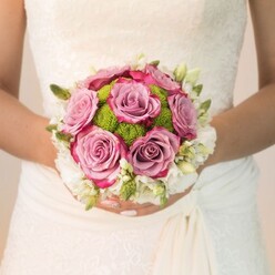A radiant moment unfolds as a bride, enveloped in a tapestry of floral lace and subtle cream drapery, holds a bouquet that could only have sprung from the heart of Epping's cherished flower shops. At its center, plush dusty mauve roses blush with hints of fuchsia-a luxurious harmony that echoes the spring sunrise filtering through High Street windows. Bright, spherical lime-green chrysanthemums cluster between the blooms, their tight texture offering a striking visual and tactile contrast, evoking the newness of life in Epping's meadows. Framing the arrangement, a delicate ring of pure white blossoms-perhaps fragrant freesias or tiny spray roses-brings a sense of clarity, their gentle scent mingling with imagined trails from the nearby forest. Intertwined foliage and green buds provide organic depth, reminiscent of a morning walk down Bell Common. The bride's hands, with nails adorned in pale pink and a hint of shimmer, provide a soft, loving cradle to this bouquet. Thoughtfully composed, this design by Flowers Epping evokes a medley of romance, hope, and quiet sophistication, perfectly suited for wedding ceremonies held in the area's rustic barns or elegant town halls. The bouquet speaks of subtle artistry and hometown pride, a symbol of enduring love blossoming in the heart of Epping.