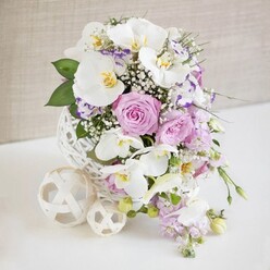 An elegant, high-end floral centrepiece is showcased against a softly textured, neutral wall, evoking the calm of a sunlit dining room in Epping on a wedding morning. At the heart of the composition sits a distinctive spherical vase, crafted from white woven strands that interlace like fine wicker or twisted paper cord, forming an intricate hollow globe. It cradles a lush bouquet of white, lavender, and blush blooms. Several large white Phalaenopsis orchids arc outward, their perfectly formed petals smooth and luminous, with warm golden and soft yellow centres that lend a hint of exotic luxury. Around them, plump roses open in shades of pastel pink and muted lilac, some fully unfurled to reveal layer upon layer of velvety petals, others still cupped, suggesting new beginnings. Intermingled are smaller, ruffled blossoms with white petals tipped in lilac, reminiscent of Lisianthus, adding a subtle frill and a painter's splash of colour. Threads of white baby's breath weave through the arrangement like fine mist, softening transitions and filling tiny gaps with a cloud-like texture. Slender stems of pale purple stock or similar trailing blooms tumble gracefully downward, giving the design a gentle cascade, while rich green foliage frames the pastels with a natural, garden-fresh feel. On the clean white tabletop sit two decorative woven orbs in a light cream tone, one large and one small, echoing the vase's open lattice and adding sculptural interest. Bathed in bright, even lighting, every detail is clearly defined, creating a refined floral display ideal for a wedding table near Epping Station or a celebratory dinner overlooking the greenery of Epping Forest.