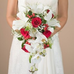 In the heart of Epping, beneath the calm light of a wedding morning, a bride in a luminous lace dress holds a magnificent cascading bouquet that gracefully defines the moment's romance. The camera catches the upper body and gentle arms of the bride, her presence blending seamlessly with the softly blurred beige background, reminiscent of the subtle tones seen near Epping Town Park. Central to the bouquet are snow-white lilies with grand, trumpet-shaped petals, standing as beacons of purity and elegance. Red roses bloom proudly amongst the lilies, their velvet petals unfurling in opulent layers, deep color evoking a sense of passion and celebration. Phalaenopsis orchids spill from the bouquet's base, their slender, graceful stems and soft ivory petals adding a sense of movement as if echoing the gentle breezes along Shooters Road. Wisps of white baby's breath and clusters of luminous red berries are artfully interwoven, offering both buoyancy and playful color reminiscent of local hedgerows. Fine, feathery greens like asparagus fern create an airy structure, surrounding each element with a lightness that feels almost magical. The composition is brought together by the bride's careful, loving hold-every flower and foliage thoughtfully selected to echo both tradition and the freshness of Flowers Epping's local style. This bouquet is not merely an accessory but a heartfelt statement, perfect for an Epping ceremony where love and elegance are celebrated under the canopy of old trees and timeless sky.