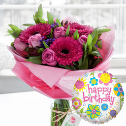 An exuberant birthday arrangement is captured in a moment of stillness, as though set on a kitchen table in Epping with daylight streaming through a nearby window. A clear glass cylindrical vase holds a structured bouquet, the stems cleanly cut and arranged with a florist's precision. Dominating the composition are bold gerbera daisies in rich fuchsia and magenta, their wide petals forming perfect circles around dark, almost black centres that draw the eye inward. Nestled between them, soft pink roses in various stages of bloom bring a more romantic note; some petals are tightly wrapped, while others are gently curling open, suggesting the slow unfolding of a celebration. Fine sprays of deep purple filler flowers weave throughout, adding depth and a touch of whimsy. Surrounding all this colour is generous, fresh greenery, each leaf catching the light and lending a garden-fresh feel that locals might associate with walks through Epping Forest on a bright spring afternoon. The stems are gathered in layers of sheer, pale pink cellophane that crinkle delicately, forming a translucent collar around the base of the blooms. A simple natural twine bow secures a small, cream-coloured gift tag with a rose emblem, giving the arrangement a handcrafted, boutique quality one might expect from an independent florist serving Epping and Loughton. Just in front and to the right, a shiny, round foil balloon completes the birthday story. Its white, lightly speckled background is scattered with colourful flower and circle motifs in yellow, blue, lime green, pink and purple, while an upbeat "happy birthday" in bold pink script dances across the centre. The softly blurred background keeps the focus on this joyful pairing, ready to surprise someone on their special day.