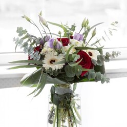 This bouquet, designed amid Epping's tranquil lanes, evokes an air of understated elegance under soft, diffused light. The arrangement is grounded by sumptuous red roses, their velvety richness providing a passionate counterpoint to the crisp, crepe-textured petals of white gerbera daisies. Each gerbera's bold brown center stands as a subtle nod to rustic charm, like the bark of ancient oaks in Epping Forest. Woven among these are lavender lisianthus, their serene hues and gently curled buds hinting at the promise of new beginnings-a fitting sentiment for anniversaries or comfort in times of reflection. Ethereal clusters of frothy white filler flowers nestle throughout, cloud-like and light, enhancing the bouquet's softness while silvery-green eucalyptus leaves arch gracefully, their rounded forms adding unique movement and a refreshing scent. The base is wrapped in broad, lush foliage, providing a polished finish that frames the bouquet with confidence and poise. Presented in a crystal-clear vase, the bouquet's vivid green stems are visible below, awash in fresh, glimmering water, testifying to Flowers Epping's commitment to quality and local freshness. This arrangement mirrors the gentle rhythm of life in Epping-unhurried, thoughtful, and elegant-delivering both visual delight and emotional warmth to kitchen tables and living rooms alike. It perfectly encapsulates the joy of sharing or receiving flowers within the welcoming spirit of our community.