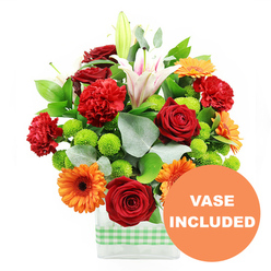 This image captures a joyful mixed-colour bouquet that feels as if it has been lifted straight from a summer garden near Epping Forest and arranged with care in a contemporary home. The composition centres on velvety deep red roses, their petals tightly cupped and romantic, alongside plush red-orange carnations whose frilled edges create a sense of movement and abundance. Radiant orange gerbera daisies, each with a defined dark centre, form bright focal points that draw the eye like bursts of sunshine. Dotted throughout the design are neat, spherical lime-green pompom chrysanthemums, adding a fresh, zesty contrast to the warmer tones and giving the arrangement a lively, almost playful rhythm. Rising gracefully above the other blooms are white Oriental lilies, some fully open to display blush-pink stripes and pollen-tipped stamens, others still in elongated buds, hinting at scent and beauty yet to unfold. The flowers are carefully placed in a clear rectangular glass vase, its straight lines and transparent sides revealing the tangle of stems and clean water, grounding the bouquet in a simple, modern style that would suit homes from Theydon Bois to the centre of Epping. Around the lower half of the vase is a light green and white gingham ribbon, tied flat like a band, evoking the relaxed charm of a country kitchen. Lush dark green leaves and sprigs of silver-green eucalyptus frame and cushion the blooms, adding depth, structure, and a subtle woodland feel. An orange circular badge with crisp white lettering reading VASE INCLUDED sits at the bottom right corner of the image, overlapping the arrangement to highlight that this floral gift comes complete and ready for same-day delivery across Epping.
