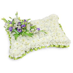 Presented on a bright white background with a soft shadow that underscores its depth, this rectangular floral pillow is lovingly built from a lavish bed of compact, snow-white chrysanthemum heads. Each bloom sits close to its neighbour, forming a uniform, velvety carpet that reads as a single, soothing surface-an image of purity and repose echoed in the smooth, sleeping-pillow shape with softly rounded edges. Tracing the entire outline is a pleated light-green ribbon trim, a carefully folded border that introduces a fresh, hopeful accent against the tranquil white. Nestled at the upper-left corner, a small but artful cluster of flowers punctuates the design: layered white lisianthus and rich purple lisianthus (some buds just unfurling) mingle with slender fern fronds and glossy foliage to create contrast, movement, and a sense of gentle reverence. The base of this accent is bound with a deeper green leaf wrap, tidily securing the stems and lending a handcrafted finish. There is a faint, clean scent suggested by the composition-green, floral, and quietly comforting, like a wreath laid near St John's Church or a tribute placed after a quiet walk through Epping Forest. As an experienced local arrangement from Flowers Epping, the pillow reads as both a respectful memorial and an expression of tender condolence, appropriate for wakes, funerals, or graveside services across Epping and the surrounding neighbourhoods.