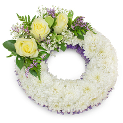 This wreath presents a quiet dignity: a perfectly round ring of small, snow-white chrysanthemums densely arranged to form a plush, comforting base that feels both immaculate and tender. Each chrysanthemum head contributes a soft, feathery texture that, when seen together, resembles a gently ruffled blanket of white petals. Tucked into the upper-left curve are three pale, creamy yellow roses, their layered petals opening like soft lanterns and symbolising care and lasting friendship. Surrounding these roses are airy clouds of white gypsophila and scattered sprays of lavender-toned statice, introducing a delicate, cool contrast that reads like the first hint of spring amid a winter palette. Interspersed lush foliage-broad, glossy leaves and finer fern fronds-adds depth, movement, and a verdant frame that makes the pale flowers sing. A discreet band of dark purple mesh or ribbon peeks from beneath the chrysanthemums at the inner and outer rims, giving the wreath an elegant edge and a thoughtful touch of colour. The scene is evenly lit with soft studio light, which highlights petal texture and casts a subtle shadow beneath the wreath on the white backdrop; the overall impression is calm, consoling and respectful. As a local florist's design often chosen for memorials in Epping, the piece carries an understated warmth appropriate for wakes, church services or a tribute laid at a graveside, offering a gentle visual expression of remembrance without ostentation.