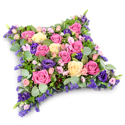 A luxurious cushion-shaped funeral tribute fills the frame, its soft, rectangular outline with rounded corners carefully sculpted from a dense mass of flowers in pink, cream, and purple. Large-headed roses in deep fuchsia, gentle blush, and buttery cream dominate the surface, many fully open to reveal their spiralling, velvety hearts. Between them, rich purple lisianthus blooms curl and twist, their layered petals creating little pockets of shadow that lend the design depth and quiet drama. Smaller spray roses in pale pink nestle close, joined by clouds of tiny lavender-toned filler flowers, much like statice or limonium, which scatter a mist of colour through the arrangement. Surrounding and threading through the blooms is a tapestry of fresh foliage: cool, silvery-green eucalyptus leaves with their smooth, rounded shapes contrast with darker, slender, fern-like greenery, adding texture and a sense of natural movement. The arrangement is photographed against a simple white background, with only a soft shadow underneath, so that every nuance of petal and leaf is clearly visible, as though the cushion has just been placed down by a caring hand. The mood is serene and contemplative, ideal for a farewell at a local Epping church or a quiet service at the nearby crematorium. This is the kind of lovingly crafted floral pillow a family might order when they want flowers to speak on their behalf, expressing sympathy, remembrance, and gentle support at a time when words feel too fragile.