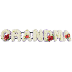 A tender, dignified floral message spells out 'GRANDMA' in large, three-dimensional letters crafted from tightly clustered white chrysanthemums that seem to glow with a soft, ivory light. Each letter is edged with a whisper of pale purple ribbon that creates a misty outline, lending depth and a peaceful rhythm to the sequence. Bright red accents - deep garden roses and rich carnations - are strategically tucked into the beginning, middle and end of the name, their saturated tones introducing warmth and heartfelt contrast to the tranquil white field. Heart-shaped red anthuriums appear at key points: the first 'A', the centre 'M' and the final 'A', their glossy surfaces reflecting light like small, steadfast hearts. The initial 'G' carries a focal cluster of deep red roses alongside a striking variegated red-and-white calla lily, the calla's sculpted form adding an elegant line to the curve.

A low-profile dark green frame supports each letter, hidden beneath foliage so the construction feels seamless and refined. As an experienced local florist serving families in Epping, from the Market Square to the quieter lanes beyond Epping Forest, I picture this tribute placed near a chapel doorway or on a sympathy display: the cool scent of chrysanthemums, the peppered sweetness of roses, and the visual steadiness of the red accents offering a quietly consoling presence. The overall effect is respectful and personalized - a measured, loving farewell to a cherished grandmother that balances tradition with intimate floral detail.