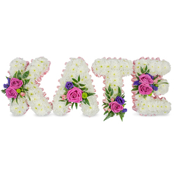 A lovingly crafted memorial display spelling 'KATE' in four separate standing letters, photographed on a clean white backdrop so every detail reads clearly. Each letter is formed from thousands of compact, snowy white chrysanthemums that create a smooth, cushiony surface; a pale pink satin ribbon delicately outlines both the outer and inner contours, adding a soft blush that frames the name with tenderness. Arranged across the letters are vivid accent clusters featuring large, velvety fuchsia-pink roses, smaller pastel pink spray roses, and fine sprays of purple or blue blooms-probably statice or small lisianthus-whose cooler tones set off the white base. Mixed foliage, from feathery fern fronds to rounded eucalyptus leaves, provides a layered, natural backdrop and suggests a faint, calming green aroma. The floral placement is meticulous: a prominent cluster sits on the diagonal of the 'K', the 'A' holds blooms near its lower right curve, the 'T' is anchored by a grouping at its base, and the 'E' is enhanced by two clusters on its middle and bottom bars-this careful choreography gives the lettering both rhythm and focus. The overall feeling is respectful and intimate, held in soft, diffuse studio light that conveys quiet dignity. As a local florist familiar with Epping and the nearby High Street and forested edges, Flowers Epping prepares these personalised sympathy tributes for wakes, funerals or remembrance services with attention to scent, texture and emotional detail.