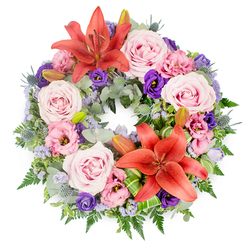 Set against a crisp white background, this elegant sympathy wreath forms a perfect, flower-packed circle, its fullness giving a sense of care and completeness. Two prominent coral-orange Asiatic lilies, fully open with gently curling petals and visible stamens, punctuate the design at opposite points, drawing the eye around the ring. Between these bold flowers, clusters of blush and light pink roses unfold in soft spirals, their velvety petals bringing a tender, almost whisper-like quality to the arrangement. Interlaced with the roses are lisianthus blooms in dusky pink and deep violet-purple, their ruffled edges catching the light and adding movement and depth. Threaded throughout, delicate sprays of lavender-blue statice create a misty, cloud-like effect, a cool contrast to the warmer tones and reminiscent of the soft, muted skies over Epping on a quiet afternoon. The underlying greenery is rich and layered: rounded, silvery eucalyptus leaves mingle with darker, glossy foliage and fine fern fronds, while spiky blue eryngium thistles contribute a subtle, textural contrast. Several unopened lily buds are nestled within the foliage, suggesting moments still to come and memories that continue to unfold. Carefully folded and striped green leaves, styled with the precision of an experienced local florist, are placed near the main lilies, adding a modern, sculptural touch. The wreath's circular shape speaks of continuity and unending love, making it a fitting tribute for a funeral or memorial service in Epping, whether it is placed by the altar of a High Street church or laid in quiet respect before a woodland-themed farewell near Epping Forest.