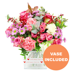 A generous, garden-fresh bouquet fills a clear square glass vase, glowing warmly against a crisp white background that makes every petal and leaf stand out, as if displayed in a bright Epping florist studio near the station. At the heart of the arrangement are plush pink roses, some in deep fuchsia and magenta, others in softer, romantic blush, their layered petals opening in spirals that suggest tenderness and care. Around them cluster ruffled carnations in coral, peach-pink, and gentle pastel hues, their soft frills creating a cloud-like texture. Nestled between these are dainty lavender and purple chrysanthemums, each bloom topped with a lively green centre that adds a zesty, playful energy to the design. Creamy white flowers are scattered throughout, providing a calm counterpoint to the brighter colours so the bouquet never feels overwhelming. Encircling the blossoms is an abundance of foliage: silvery-green eucalyptus with rounded leaves, deeper green sprigs, and a hint of reddish-brown foliage that lends an almost autumnal warmth, reminiscent of walks along Epping Forest's leafy paths. The stems are clearly visible through the transparent vase, which is wrapped at the base with a smooth white band printed with small red hearts and the affectionate phrase love all my love, subtly suggesting this gift is meant for someone treasured. In the bottom right corner of the image, an orange circular graphic announces VASE INCLUDED in bold white capitals, making it easy to imagine this bouquet arriving as a complete, ready-to-enjoy gift for homes around Theydon Bois, Coopersale, or central Epping. The overall feeling is one of thoughtful, uplifting affection, ideal for Valentine's Day, Mother's Day, or simply brightening a loved one's week.
