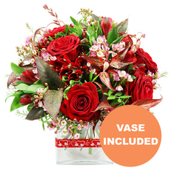 A richly romantic floral display is captured against a clean white backdrop, drawing the eye first to a cluster of sumptuous red roses that appear almost velvety to the touch. Some roses are tightly furled, their petals wrapped close like a secret, while others are more open, revealing swirling hearts that hint at deep affection and timeless love. Threaded between them are smaller, equally intense crimson flowers evocative of alstroemeria and carnations, their multiple petals and speckled throats adding movement and complexity to the dome-shaped bouquet. Delicate sprays of pale pink and white waxflower drift throughout the arrangement, their miniature cup-shaped blossoms and rounded buds softening the bold reds with a gentle, cloud-like texture. Around the blooms, a generous collar of lush greenery spreads outward: glossy dark green leaves, finer fresh green tips, and distinctive burgundy and copper-toned foliage with subtle veining that echoes the richness of the roses and introduces a note of refined drama. The bouquet sits neatly in a modern, clear rectangular glass vase, its straight lines contrasting beautifully with the organic curves of the flowers and making it an easy piece to place on a dining table in an Epping home, perhaps overlooking the greenery near Bell Common. A festive red ribbon dotted with white heart outlines encircles the vase, a playful nod to romantic celebrations or thoughtful gestures. In the lower right corner of the picture, an orange circular label featuring crisp white capital letters clearly states "VASE INCLUDED," signalling that this arrangement arrives as a complete, ready-to-enjoy gift for local flower delivery around Epping.