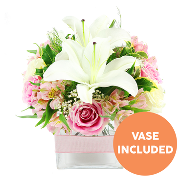 Set against a crisp white backdrop, this polished floral arrangement combines classic romance with modern simplicity, much like the mix of old and new you find strolling through Epping's High Street. Two pristine white Oriental lilies dominate the centre, their generous petals opening into star-shaped blooms, each one cradling rich reddish-brown stamens that draw the eye inward. Around them, soft pink roses in different stages of bloom-from tight buds with swirling centres to fully unfurled blossoms-create a sense of movement and unfolding emotion. Threaded delicately between these are pink and white alstroemeria, some petals kissed with fine yellow speckles that echo early morning sunlight. Light, cloud-like sprays of white baby's breath soften the edges and add a gentle, romantic haze. The flowers rise from a clear, low square glass vase, where the stems are visible through the water, giving the design a crisp, contemporary edge well suited to a calm living room near Stonards Hill or a cosy kitchen by Epping station. A pale pink satin ribbon encircles the middle of the vase, tied neatly to introduce a hint of gift-wrap charm without overwhelming the clean lines. Rich greenery in various shapes and textures-broad shining leaves, narrower blades, and feathery fronds-frames the blooms and deepens the contrast between the white and pink tones. In the bottom right corner, a bright orange circular label with bold white text announces VASE INCLUDED, clearly indicating that the glass container is part of the arrangement. The bouquet feels poised and serene, perfect for conveying gentle congratulations, get-well wishes, or quiet celebrations delivered across Epping.