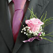 This image offers an intimate view of formalwear prepared for a special celebration in Epping, perhaps a wedding at a local church or a prom evening gathering near Epping Station. The frame captures only the upper torso of a person, drawing attention to the interplay between tailored style and careful floristry. A deep charcoal, almost black suit jacket forms a refined backdrop, its lapel clean and crisp. Underneath, a pale, finely checked dress shirt adds a discreet pattern, while a luminous fuchsia or magenta tie, smooth and silky, runs straight down the centre like a ribbon of colour. Pinned to the left lapel is a thoughtfully crafted boutonniere that feels both modern and romantic. At its heart, a single pastel pink rose opens in a perfect spiral, with petals that deepen to a richer pink at the centre before fading to a soft blush at the edges. Surrounding the rose, delicate sprays of cloudlike baby's breath cluster in tiny white blooms, softening the contrast against the dark suit. Behind them, slender green shoots of decorative foliage arc gently upwards, suggesting movement and the natural ease of a garden border along a quiet Epping lane. The base of the arrangement is carefully bound with deep green wrapping and finished with a small, satiny purple bow, positioned neatly above the breast pocket and subtly catching the light. The colours of the ribbon and rose harmonise beautifully with the bold tie, giving a sense of coordinated thoughtfulness. Everything about the image suggests fresh flowers prepared on the day, perhaps by a local florist who understands how small details help a groom or guest feel truly ready for a cherished occasion in the heart of Epping.