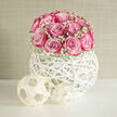 This image shows a carefully composed floral design where a dome of lush pink roses becomes the immediate focal point, each bloom tinted from pale blush at the outer petals to a deeper, more confident fuchsia at the centre. The roses are closely clustered, creating a luxurious cushion of colour that feels both modern and romantic. Encircling them is a delicate band of white gypsophila, the baby's breath forming a misty, almost cloud-like fringe that softens the edges of the arrangement. All of this sits in a striking white spherical vessel, hand-woven in an open lattice that loops and curls like an abstract sculpture. The orb's airy design allows light to pass through, so you see intriguing patterns of brightness and shadow on the surface below, much like sunlight dappling a path in one of Epping's quiet residential streets. The overall palette is crisp and light: creamy whites, fresh pinks, and subtle shadows, suggesting a calm, sophisticated setting-perhaps the centre of a wedding table at a country house near Epping, or a showpiece on a hallway console as guests arrive. As an experienced local florist, you can almost feel the contrast between the smooth, cool wicker of the globe and the silky softness of the petals, and sense the gentle rose perfume that would fill a room. The design speaks of joyful celebration and thoughtful design, ideal for couples who want their Epping wedding flowers to blend contemporary styling with a tender, romantic mood.