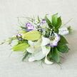 This bouquet rests on a neutral, textured fabric, its horizontal composition emphasizing a graceful sweep of flowers and foliage. The white Oriental lilies take centre stage, each petal smooth and luminous as cream, some open to display warm yellow stamens dotted with dark pollen, others tight green buds that promise unfolding drama. Surrounding them, lisianthus blooms show a soft white heart that graduates into lilac and rich purple ruffles at their edges, giving a watercolor effect across the petals. Sprigs of delicate Baby's Breath are woven through the arrangement, their tiny white florets creating a soft haze that offsets the bolder blooms. The greenery anchors the grouping with a range of textures: glossy, broad leaves lay a deep verdant foundation while slender, almost grassy spikes and feathery fronds introduce vertical lines and airy gaps. A white satin ribbon wraps the stems in a clean, bridal finish; discreet pearl pins or small sparkling studs secure the wrap and add a refined, celebratory flicker. The scent is subtle - an almost spicy lift from the lilies balanced by the sweet, tender perfume of eustoma - conjuring images of early light over Epping Forest or a quiet moment on the High Street. Arranged with thoughtful asymmetry and fresh materials, this bouquet suits weddings, anniversary gestures, or elegant home centrepieces, and it reflects the measured, seasonal sensibility of florists who deliver across Epping and nearby villages.