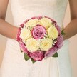 In a scene filled with romance and quiet anticipation, the camera captures an Epping bride holding a sumptuous, hand-tied bouquet in the glow of pure, natural light. The bouquet, rounded and lush, brings together creamy, lemon-tinged yellow roses mingled harmoniously with vibrant fuchsia pink blooms and softer mauve-pink highlights-petals whose hues seem to echo the garden landscapes of Epping Forest on a spring morning. Ethereal wisps of white Gypsophila are threaded within the roses, giving the arrangement a whisper-light airiness that dances in the light, while dark green leaves suggest the surrounding Epping countryside. The bride's bare forearms, arms encircling the bouquet at her waist, convey nurturing care and excitement, her delicate touch giving voice to the tender intimacy of local wedding celebrations. Soft, intricate lacework adorns the bodice of her pale ivory gown, and the fluid fabric below gently frames the vibrant flowers, capturing the classic yet modern taste found among Epping's creative community. Every petal and sprig is illuminated by bright, even daylight, which accentuates the bouquet's freshness and the shimmering details of silk and lace. The blurred, pale background lets the passion and expertise of Epping's florists shine, evoking the joy and serenity of a community wedding close to Epping's beloved green spaces and village halls. This evocative image brings to life not only the artistry of local floral design but also the love and anticipation that makes each wedding day uniquely treasured.