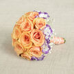 Laid horizontally on a woven light beige fabric that resembles linen, this bridal bouquet is photographed from a slightly raised vantage so the viewer can appreciate both the rounded form and the delicate details. The centre is a dense cluster of peach and soft orange roses, each blossom opening in concentric curls that create a warm, sumptuous texture and catch the light like a soft glow. Sprinkled through the arrangement are white lisianthus flowers, their ruffled petals edged in a striking violet - small flashes of cool colour that lift and modernise the otherwise classic palette. Tiny stems of baby's breath weave through the massed blooms, giving pockets of air and a whispery, whimsical cadence between heavier petals. The handle is neatly wrapped in a pale peach satin ribbon, its smooth surface contrasted by the lustre of an off-white pearl strand wound in an elegant spiral, a tactile flourish that suggests tradition and quiet luxury. The soft, natural lighting emphasizes petal translucency and the satin sheen while leaving gentle shadowing to accentuate depth. One can almost sense the fresh rose fragrance mingling with the green, earthy note of stems - a sensory impression that pairs well with celebrations held near St John's Road or a ceremony in a small Epping chapel. The bouquet reads as lovingly composed and restrainedly romantic, suitable for brides who want a mix of classic warmth and a tasteful modern edge, and it feels like a thoughtful keepsake after the day is done.