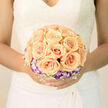 Here the composition reads like gentle poetry, a bridal bouquet designed to sit quietly at the centre of a wedding photograph. Twenty or so apricot roses are arranged in a symmetrical, gathered cluster, each bloom unfurling with creamy petals that glow with warm, peachy light. Surrounding that central mass is a crown of purple-and-white lisianthus whose cool tones bring a twilight-like hush to the palette, as if peach sunlight has met soft evening clouds. Interwoven through the blooms, a delicate lattice of white baby's breath and airy fillers softens edges and lends a feather-light texture that makes the whole bouquet seem to float when lifted.
The surface of each petal shows gentle veins and the faintest satiny sheen where the light brushes across it; close observation reveals the florist's exacting hand in the precise placement and balance. I imagine this bouquet resting against a bride's lace gown on Epping High Street outside the tube station or just beyond the trees at the edge of Epping Forest-timeless, understated, and utterly refined. The scent would be quietly sweet, the tactile contrast of velvety petals and brittle lace warmly familiar. It's an arrangement meant for ceremony and photographs, crafted to sit beautifully in a bride's hands while offering a soft narrative of hope, promise, and the intimate joy of a wedding day.