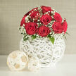 A lush cluster of red roses forms a near-perfect dome at the heart of this image, each flower at the ideal stage of bloom: petals unfurling in neat, velvety spirals, rich with the promise of scent and softness. Between the crimson heads, delicate stems of white baby's breath weave in and out, their tiny, cloud-like blossoms adding a lacy border that frames the roses and tempers their intensity. A few deep green leaves hide in the shadows of the arrangement, offering small flashes of foliage that remind you these blooms were freshly cut. The bouquet is nestled into a distinctive spherical vase, crafted from smooth, white, wicker-like strands that cross and loop around each other in an open, airy pattern. Light filters through the gaps in the weave, creating subtle highlights and shadows that ripple across the clean white tabletop below. To one side, two decorative orbs made from pale, natural fibre sit quietly, mirroring the texture and curves of the vase while introducing softer cream tones. Behind, a neutral wall in light beige or grey fades into the background, gently textured but visually calm, giving centre stage to the vivid reds and crisp whites. The arrangement feels like it belongs in an elegant Epping home, perhaps in a sunlit room overlooking the green spaces that lead toward Theydon Bois or the edge of Epping Forest. It suggests the kind of romantic yet contemporary design chosen for wedding receptions, vow renewals, or special anniversary dinners hosted in local Epping venues, where every detail is considered to create a timeless, welcoming atmosphere.