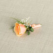In soft natural light this peach rose boutonniere is presented with the attentive restraint of a seasoned local florist: its single bloom is a study in gentle colour, from a warm apricot heart to pale peach edges, petals layered like a shy promise. Sprays of gypsophila float around the rose like tiny stars, their delicate white dots bringing a dreamy, whimsical counterpoint to the rose's plush surface. Verdant loops of foliage encircle the base, their glossy texture and crisp curves grounding the arrangement and offering a cool, herbal contrast to the flower's velvety warmth. The stem is wrapped with satin ribbon in a matching peach tone, tied with a neat, low-profile knot whose subtle sheen catches glints of light - a finishing touch that speaks to careful technique and an eye for proportion. You can almost sense the faint sweet scent of the rose and the airy perfume of summer mornings beneath the canopy of Epping Forest-a local scene where many couples choose to exchange vows - and it's the sort of boutonniere a bridegroom might slip into his lapel before stepping out on the High Street for photographs by the red-brick clock or St. John's church. As created by Flowers Epping, this piece feels simultaneously tender and enduring: modest in scale yet precise in execution, designed to complement formalwear without overwhelming it, and to become a subtle, remembered token in hugs, pictures, and quiet moments on a wedding day.