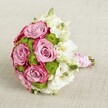 A luxurious hand-tied bridal bouquet rests on a softly textured light beige fabric, photographed from a gently elevated angle so you can clearly sense its rounded, compact shape. At the centre, several large roses open fully in a romantic two-tone of soft lavender-pink fading into a deeper fuchsia pink at the petal edges, the kind of nuanced colour you often see in premium wedding flowers chosen by brides in Epping. Between these velvety blooms, small chartreuse green button chrysanthemums peek through, their tight, domed heads adding a crisp, almost zesty texture that makes the pinks appear even richer. Delicate white lisianthus, with layered petals and tiny greenish-yellow centres, weave through the design like soft clouds, while a few unopened buds signal the bouquet's freshness, as if it has just been prepared for same-day delivery to a ceremony near Epping Forest. The stems are neatly gathered into a tidy handle, wrapped in a smooth pink satin ribbon that catches the light with a muted sheen. Spiral around this ribbon is a strand of tiny white pearls, creating a refined, almost heirloom feel that would suit a classic church wedding in the heart of Epping or an intimate celebration at a local country venue. The neutral background allows the interplay of pink, green, and white to stand out, evoking romance, joy, and careful craftsmanship. You can almost imagine a bride lifting this bouquet, the satin cool against her hand and the scent of fresh roses surrounding her as she walks down the aisle.