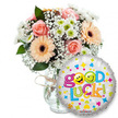 A vibrant and encouraging gift set captured against a clean white backdrop: on the left, a generous bouquet sits in a clear glass vase, its base tied with a simple twine bow, and to the right a round, silver metallic foil balloon leans forward, overlapping the lower portion of the stems. The arrangement is an intimate study in soft, comforting tones - powder-pink and pale peach roses with tightly furled petals nestle among peachy-orange Gerbera daisies whose dark brown centers give a warm focal point. Sprays of delicate white baby's breath thread through the bouquet like lace, while clusters of small white and lime-green chrysanthemum florets add a bright, crisp texture. Deep green foliage peeks from between blooms, lending depth and a cool leafy contrast to the warm peach and pink palette. The balloon's reflective silver surface is freckled with cheerful confetti shapes - blue and pink hearts, yellow and blue stars, tiny orange and green crosses - and the message Good Luck! arcs across it in playful, multi-coloured letters; two vivid yellow smiley faces replace the o's in Good, one winking, one playfully sticking out its tongue, giving the whole composition a lighthearted, encouraging personality. Soft, neutral lighting emphasizes petal texture and the balloon's sheen, while the simple vase keeps attention on the flowers and message. This ensemble reads as a thoughtful send-off or a brave-wish for exams, interviews, graduations or new beginnings - a tactile, fragrant token of encouragement designed to bring colour and hope into someone's day.