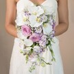 This elegant bridal portrait captures a bride from Epping, pictured from mid-chest downward, her bare arms encircling a lavish, shower-style cascading bouquet that steals the scene. Set before a softly blurred, beige background reminiscent of early light over Epping's historic streets, the bouquet spills downward in waves, entwining pristine white phalaenopsis orchids, plush blush and soft pink garden roses, and light lavender-toned lisianthus. The orchids' rich, yellow-touched centres draw the eye, their broad petals feeling almost silken, while roses unfold in soft, romantic fullness, evoking the classic charm found in gardens along Bell Common. Sprays of baby's breath and slender strands of green foliage add a dreamy softness, filling out the bouquet without overcomplicating its shape. The bride's strapless white gown, adorned with detailed lace at the bodice, subtly glows in the diffused light, focusing all attention on the exquisite florals. Every element has been chosen for harmony and texture-a composition that captures the spirit of celebration in Epping. This showstopping bouquet, crafted by skilled local florists, is especially favored for weddings at nearby historic venues, offering a sense of timeless romance while embracing the gentle luxuries of Essex floral design.