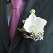 A single white orchid, luminous and poised, forms the heart of this boutonniere, resting just above the pocket of a charcoal suit. Its petals are broad and gently cupped, almost porcelain-smooth, catching the light in a soft, velvety sheen. At the centre, a subtle blush of ivory draws you inward, surrounded by a collar of cool, silvery-green leaves that frame the bloom with understated elegance. Sprigs of lilac-toned flowers and tiny white gypsophila weave between the foliage, like small wildflowers gathered from the edges of Epping Forest, bringing a sense of freshness and natural grace. The stems are bound with a narrow violet ribbon, its satiny texture glimmering against the darker jacket, and its colour thoughtfully matched to the purple-pink tie beneath. The ribbon is tied with a careful, almost invisible precision, allowing the flowers to appear as though they are simply resting there, effortless yet meticulously crafted. The contrast between the crisp orchid and the rich fabric of the suit gives the boutonniere a floating, ethereal quality, as if it were suspended in the soft daylight often seen over Epping High Street on a clear morning. This floral accent feels ideal for a wedding, anniversary, or formal gathering held in and around Epping, when every detail is chosen to reflect love, personality, and quiet sophistication. It carries the mood of anticipation-a moment just before vows are spoken or a toast is raised-capturing in flowers the tender excitement of a day that matters.