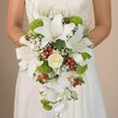A close-up image shows a bride in a flowing white dress, holding an exceptional teardrop bridal bouquet evocative of Epping's romantic woodlands and vibrant garden tradition. The bouquet is abundant, its structure characterised by bold, gleaming white lilies with trumpet forms and subtle, cinnamon freckles, giving a sense of grandeur and purity. Resting at its centre, a single creamy white rose unspools layers of petals, softly scented and timeless-much like those discovered in tranquil corners of Stonards Hill's flourishing gardens. Lush, lime-green mums are interspersed throughout, lending modern flair and a crisp visual counterpoint to the classic whites, while airy clusters of gypsophila cloud the arrangement with their gentle, milky-white texture. The bouquet is joyously peppered with glossy bright red Hypericum berries, their playful pop conjuring the festive spirit found in Epping's seasonal flower stalls. Cascading downwards, elegant white Phalaenopsis orchids create a waterfall effect, their blooms fluttering with graceful motion and imparting a touch of effortless luxury. The bride's hands, partly veiled by the subtle lace details of her gown, gently cradle this masterpiece. Wisps of fine green foliage peek out from beneath the flowers, echoing the natural serenity of local green spaces. Bathed in natural, soft light, this bouquet is an ideal choice for anyone in Epping seeking a harmonious blend of traditional bridal beauty and lush, contemporary detail for their special ceremony.